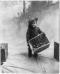 A small boy carries a basket of apples while holding an apple in his mouth, 1911